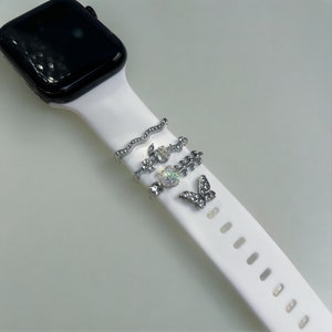 Pode incluir: Uma pulseira branca de Apple Watch com pingentes prateados. Os pingentes incluem uma linha ondulada, uma flor, uma folha e uma borboleta.