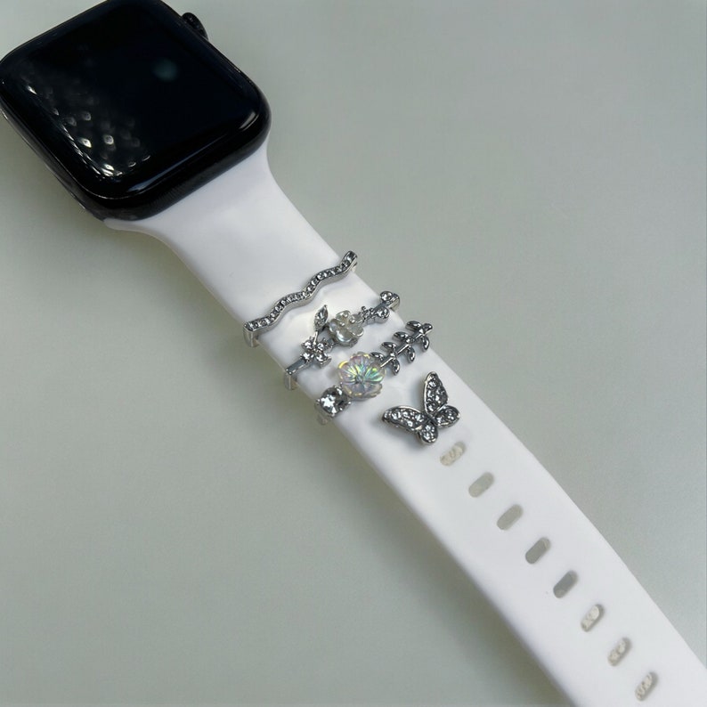 Pode incluir: Uma pulseira branca de Apple Watch com pingentes prateados. Os pingentes incluem uma linha ondulada, uma flor, uma folha e uma borboleta.