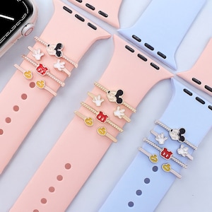 May include: A set of four gold-coloured watch band charms featuring Mickey Mouse, his gloves, and a red circle. The charms are shown on two different coloured watch bands, one pink and one light blue.