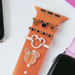 K&ouml;nnte beinhalten: Orangefarbenes Silikonarmband f&uuml;r die Apple Watch mit drei goldfarbenen Anh&auml;ngern: einem K&uuml;rbis, einem Mickey-Maus-Kopf und einem mit Strass besetzten Mickey-Maus-Kopf.