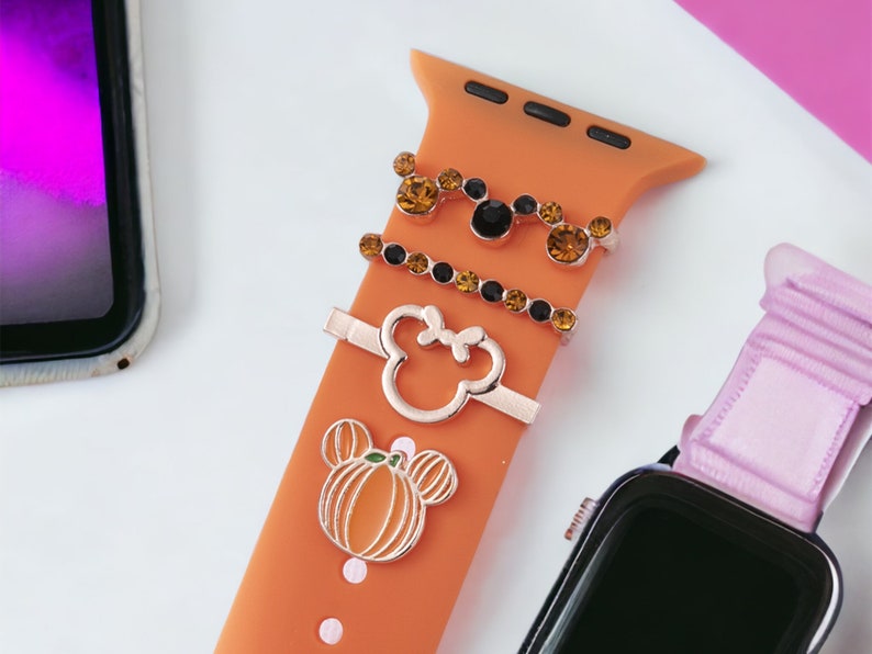K&ouml;nnte beinhalten: Orangefarbenes Silikonarmband f&uuml;r die Apple Watch mit drei goldfarbenen Anh&auml;ngern: einem K&uuml;rbis, einem Mickey-Maus-Kopf und einem mit Strass besetzten Mickey-Maus-Kopf.