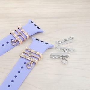Puede incluir: Dos correas de reloj de silicona púrpura con dijes decorativos. Los dijes son de metal y tienen un diseño de corazón con diamantes de imitación transparentes. Los dijes están unidos a la correa del reloj con un lazo.
