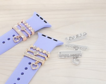 May include: Two purple silicone watch bands with decorative charms. The charms are made of metal and feature a heart design with clear rhinestones. The charms are attached to the watch band with a loop.