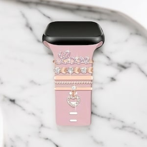 Puede incluir: Una correa de reloj de silicona rosa con una esfera negra. La correa está decorada con varios abalorios de oro rosa, incluyendo un abalorio en forma de corazón con pedrería, un abalorio de cadena y un abalorio que dice "Love" con pedrería.