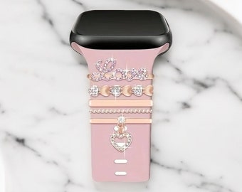 May include: A pink silicone watch band with a black watch face. The band is decorated with several rose gold charms, including a heart charm with rhinestones, a chain charm, and a charm that says "Love" with rhinestones.