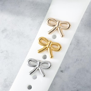 May include: Three gold-toned bow-shaped charms in different sizes. The charms are on a white surface.