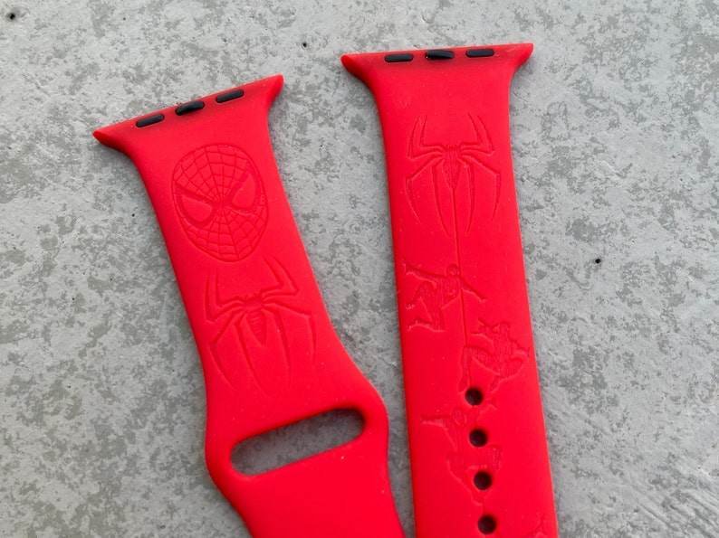 May include: Two red silicone Apple Watch bands with a Spider-Man design. One band features a close-up of Spider-Man's face and the other band features Spider-Man swinging through the city.