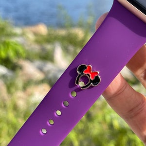 Puede incluir: Una correa de reloj morada con un dije decorativo con un diseño de orejas de Minnie Mouse negras y rojas. El dije es de color dorado y está sujeto a la correa. El fondo está borroso, mostrando vegetación y agua.
