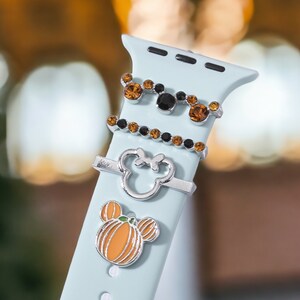 K&ouml;nnte beinhalten: Ein hellblaues Apple Watch Armband mit drei silbernen Charms. Die Charms sind ein K&uuml;rbis, ein Mickey Mouse Kopf und ein Mickey Mouse Kopf mit orangefarbenen und schwarzen Strasssteinen.