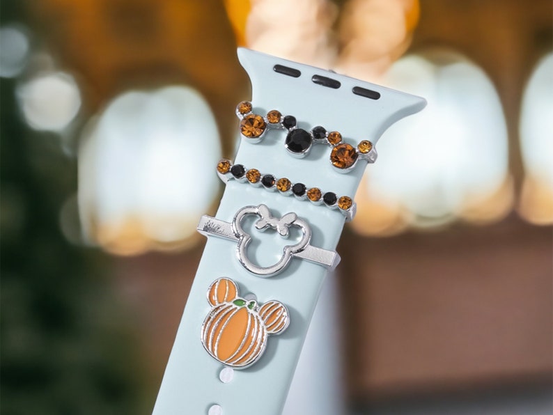 K&ouml;nnte beinhalten: Ein hellblaues Apple Watch Armband mit drei silbernen Charms. Die Charms sind ein K&uuml;rbis, ein Mickey Mouse Kopf und ein Mickey Mouse Kopf mit orangefarbenen und schwarzen Strasssteinen.
