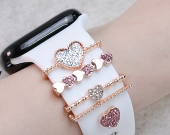 May include: A white Apple Watch band with four rose gold heart-shaped charms. The charms are adorned with sparkling rhinestones.