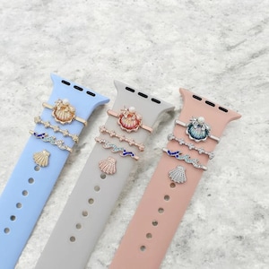 May include: Three Apple Watch bands in blue, gray, and pink with a set of five charms on each band. The charms are a seashell, a wave, and three different styles of seashells with pearls.
