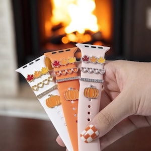 May include: Three Apple Watch bands with fall-themed charms. The bands are white, orange, and pink. The charms include pumpkins, leaves, and hearts.