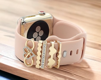 May include: A pink silicone Apple Watch band with gold-toned metal charms. The charms include a heart with an infinity symbol, a row of small diamonds, and a wavy design.