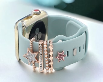 May include: A light blue silicone Apple Watch band with a gold watch face and four decorative charms. The charms are a star with rhinestones, a bar with rhinestones, a pearl chain, and a pink star with rhinestones.