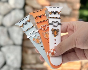 Pode incluir: Três pulseiras para Apple Watch com diferentes pulseiras de silicone coloridas e pingentes. Os pingentes são uma abóbora, uma cabeça do Mickey Mouse e uma cabeça do Mickey Mouse preta com pedras. As pulseiras são azul claro, laranja e branco.