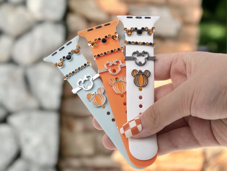 K&ouml;nnte beinhalten: Drei Apple Watch-Armb&auml;nder mit verschiedenen farbigen Silikonb&auml;ndern und Charms. Die Charms sind ein K&uuml;rbis, ein Mickey-Maus-Kopf und ein schwarzer Mickey-Maus-Kopf mit Strasssteinen. Die Armb&auml;nder sind hellblau, orange und wei&szlig;.