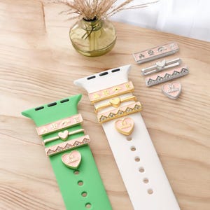 Könnte beinhalten: Eine Sammlung dekorativer Charms für Smartwatches. Die Charms sind in verschiedenen Designs erhältlich, darunter Herzen und geometrische Muster, mit rosa und goldenen Akzenten. Die Charms werden auf grünen und weißen Armbändern präsentiert.