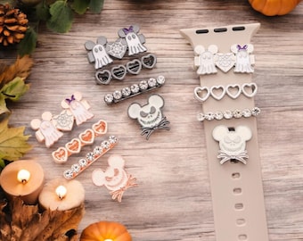 Könnte beinhalten: Eine Sammlung dekorativer Charms für ein Smartwatch-Armband mit Halloween- und Disney-Motiven. Die Charms zeigen Geister, Herzen und mausförmige Charaktere mit Strassakzenten. Die Farbpalette umfasst Weiß, Schwarz und Roségold.
