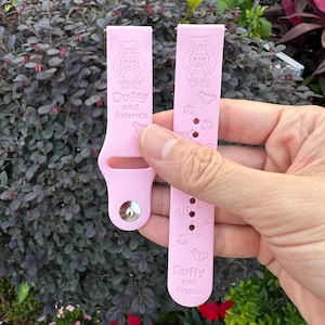 May include: Two light pink watch bands with the text "Duffy and Friends" and cartoon character designs. One band has a buckle, and the other has several holes. The bands are held up against a background of green and purple foliage.