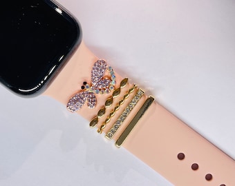 May include: A pink Apple Watch band with a butterfly charm and several gold-colored decorative bands. The butterfly charm is adorned with pink and iridescent crystals. The watch face is black.