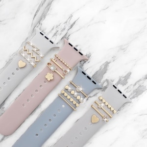 Puede incluir: Cuatro correas de Apple Watch coloridas en gris, rosa y azul, adornadas con abalorios decorativos. Los abalorios incluyen corazones, llaves y formas geométricas, algunas con pedrería. Presentado sobre una superficie de mármol.