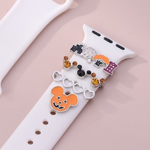 May include: A white Apple Watch band with a variety of charms attached. The charms include a pumpkin-faced Mickey Mouse, a heart charm, a Mickey Mouse head, a charm with a castle and a sphere, and a charm with a building.