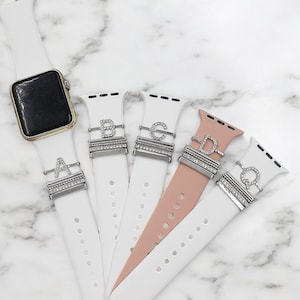 Puede incluir: Cinco bandas diferentes para Apple Watch con dijes intercambiables que presentan las letras A, B, C, D y Q. Los dijes están hechos de metal plateado y tienen diamantes de imitación transparentes.
