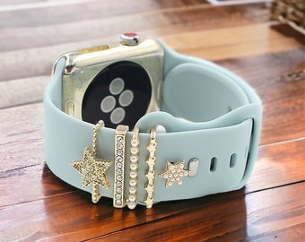 May include: A light blue silicone Apple Watch band with gold-toned charms. The charms include a star with rhinestones, a bar charm with rhinestones, a pearl and star charm, and a star charm with pearls.