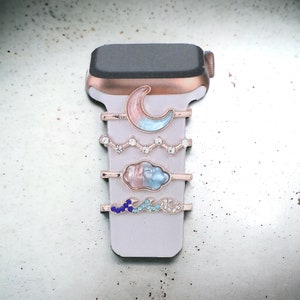 Puede incluir: Un conjunto de cinco correas de reloj con diferentes encantos. Los encantos incluyen una luna creciente, una nube y una ola. Las correas son todas de color oro rosa y tienen diamantes de imitación transparentes.