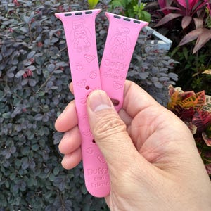 May include: Two pink watch bands with a cartoon character design. The bands feature the text "Duffy and Friends" and various character illustrations. The bands are held in a hand against a background of green and red foliage.