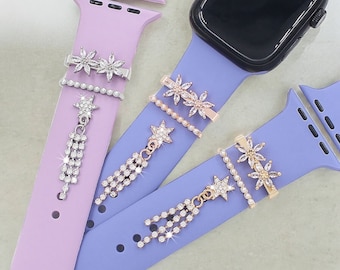 May include: Two Apple Watch bands with decorative charms. The bands are purple and light blue. The charms are silver and gold with crystal embellishments. The charms are shaped like stars and flowers.