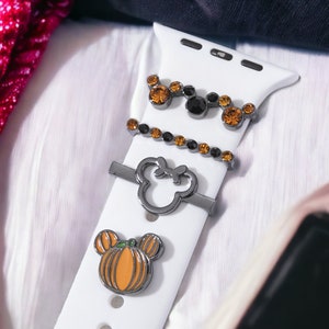 K&ouml;nnte beinhalten: Ein wei&szlig;es Apple Watch Armband mit drei Charms: ein K&uuml;rbis-f&ouml;rmiger Mickey Mouse Kopf, ein Mickey Mouse Kopf mit schwarzen Strasssteinen und eine Reihe schwarzer und orangefarbener Strasssteine.