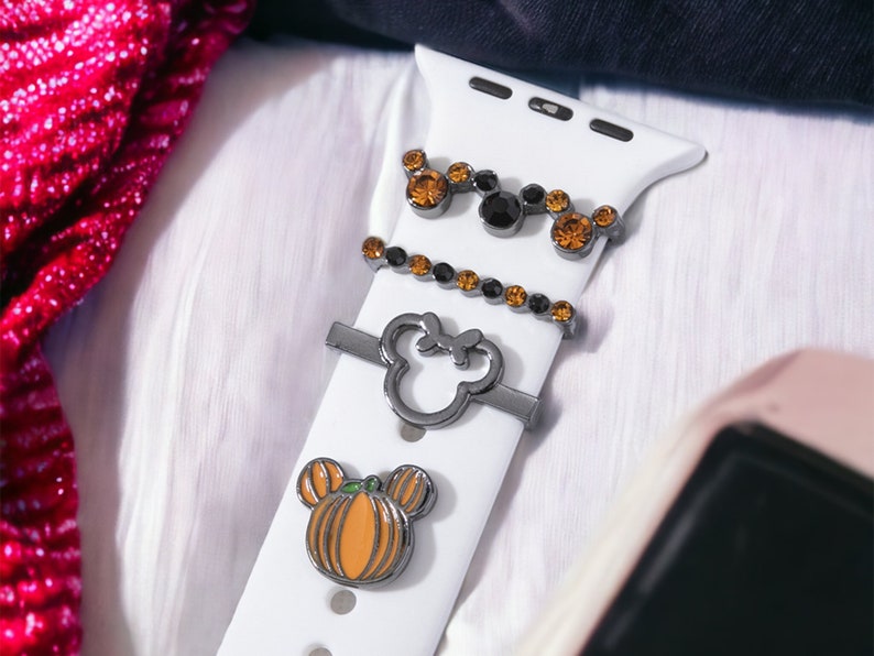 K&ouml;nnte beinhalten: Ein wei&szlig;es Apple Watch Armband mit drei Charms: ein K&uuml;rbis-f&ouml;rmiger Mickey Mouse Kopf, ein Mickey Mouse Kopf mit schwarzen Strasssteinen und eine Reihe schwarzer und orangefarbener Strasssteine.