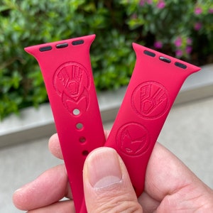 May include: Two red silicone Apple Watch bands with a raised design of a superhero's hand and symbol. The bands are shown on a person's wrist.