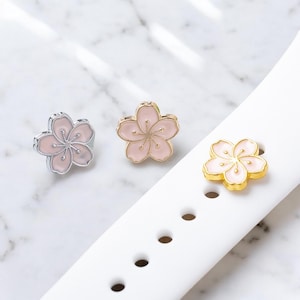 May include: Three pink and gold cherry blossom flower charms. The charms are on a white watch strap.