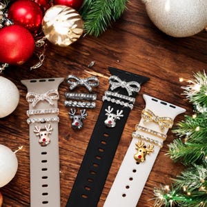 Puede incluir: Tres correas de reloj en gris, negro y blanco, adornadas con abalorios festivos. Cada correa presenta un lazo, un anillo de pedrería y un dije de reno. El fondo incluye adornos navideños y vegetación, creando un tema festivo.