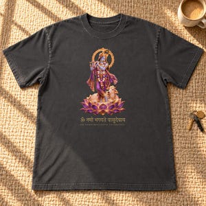 May include: A charcoal gray short-sleeve t-shirt featuring a graphic of Lord Krishna playing a flute while standing on a lotus flower next to a cow. Below the graphic is the Sanskrit text "ॐ नमो भगवते वासुदेवाय" and the English transliteration.