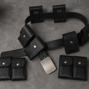 Pode incluir: Um cinto utilitário preto com uma fivela metálica retangular lisa e vários bolsos destacáveis. Cada bolso é fixado com um fecho de botão de pressão prateado. O cinto é feito de um material texturizado com costuras reforçadas.