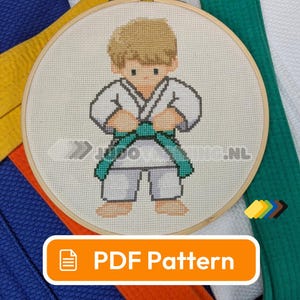 Op de afbeelding: Kruissteekborduurwerk van een jonge judoka in een witte gi met een groene band. Het ontwerp is geplaatst in een houten hoepel, met de tekst "PDF Pattern" in een oranje kader. Judo-trainingsuitrusting is zichtbaar op de achtergrond.