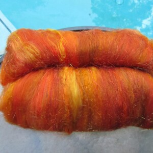 May include: A rectangular batt of hand-dyed wool roving featuring a vibrant blend of fiery orange, deep red, and bright yellow fibers. The texture is soft and fibrous, with a slight sheen throughout the material, showcasing a variegated color pattern.