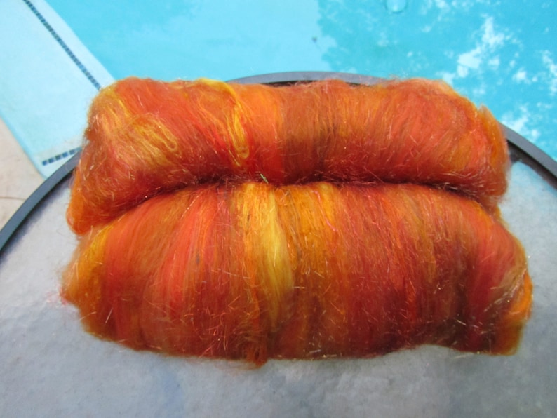 May include: A rectangular batt of hand-dyed wool roving featuring a vibrant blend of fiery orange, deep red, and bright yellow fibers. The texture is soft and fibrous, with a slight sheen throughout the material, showcasing a variegated color pattern.