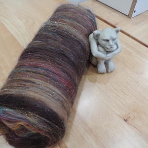 May include: A large roll of brown yarn with strands of silver and other colors. A small, light-colored statue of a seated lion-like creature is next to the yarn on a wooden surface. The yarn has a soft, textured appearance.