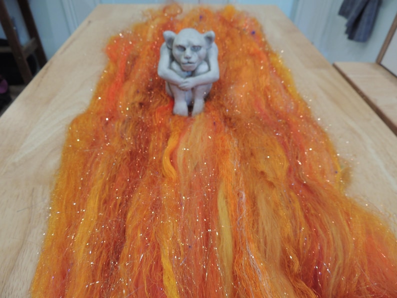 May include: A small, light gray gargoyle figurine in a seated, hunched position, placed atop a long, flowing pile of vibrant orange, yellow, and red wool roving fibers that feature scattered, shimmering metallic glitter throughout the material.