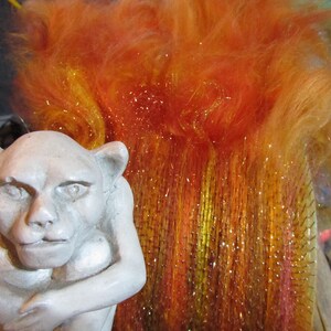 May include: A light gray stone-colored gargoyle figurine with pointed ears and a hunched posture positioned next to a rectangular carding brush filled with vibrant orange, yellow, and red synthetic fibers that have a metallic, shimmering finish.
