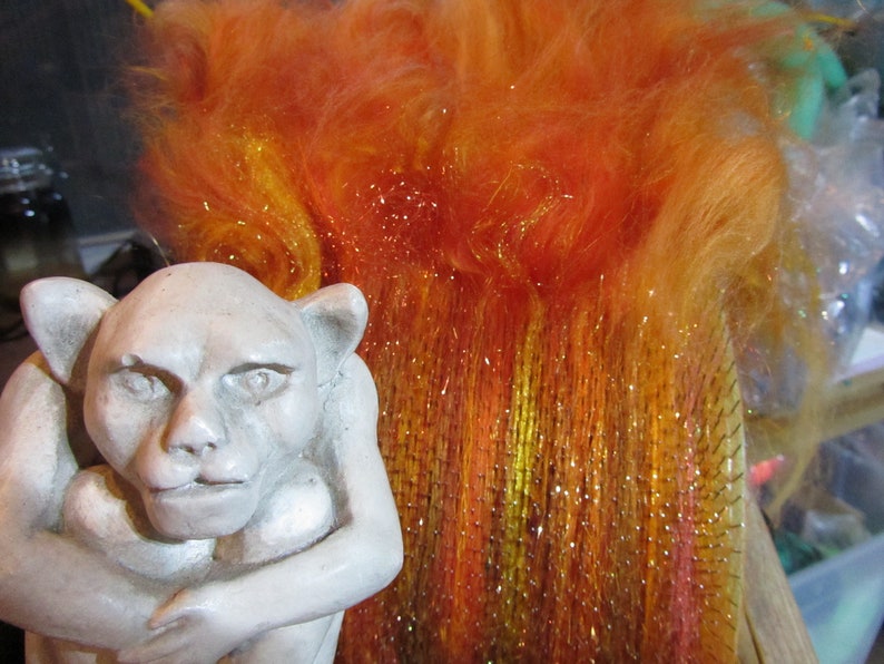 May include: A light gray stone-colored gargoyle figurine with pointed ears and a hunched posture positioned next to a rectangular carding brush filled with vibrant orange, yellow, and red synthetic fibers that have a metallic, shimmering finish.