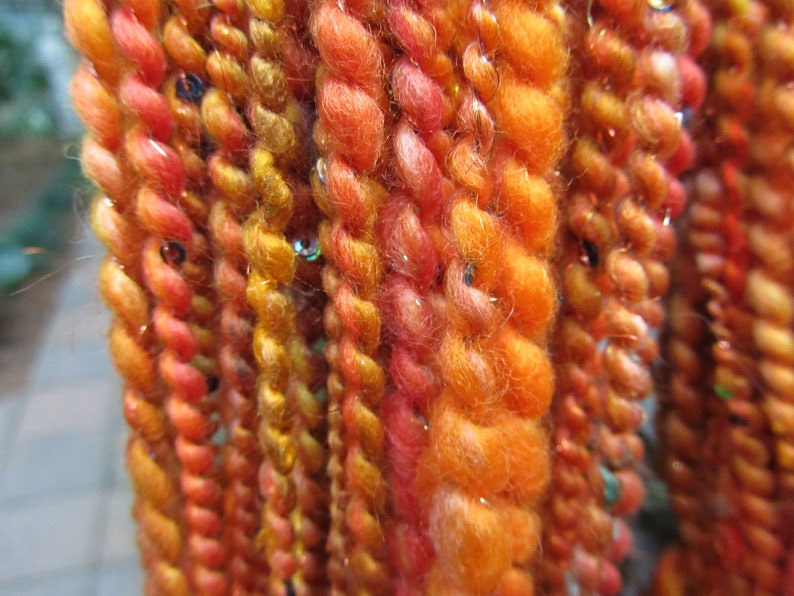 May include: A collection of multi-textured yarn strands in vibrant shades of orange, yellow, and red. The yarn features a twisted construction with integrated metallic gold threads and small, reflective sequins scattered throughout the fibers.