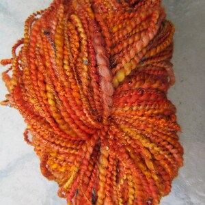 May include: A skein of textured art yarn in vibrant shades of orange, red, and yellow. The yarn features a multi-ply construction with varying thicknesses, incorporating metallic threads and small reflective sequins throughout the twisted fibers.