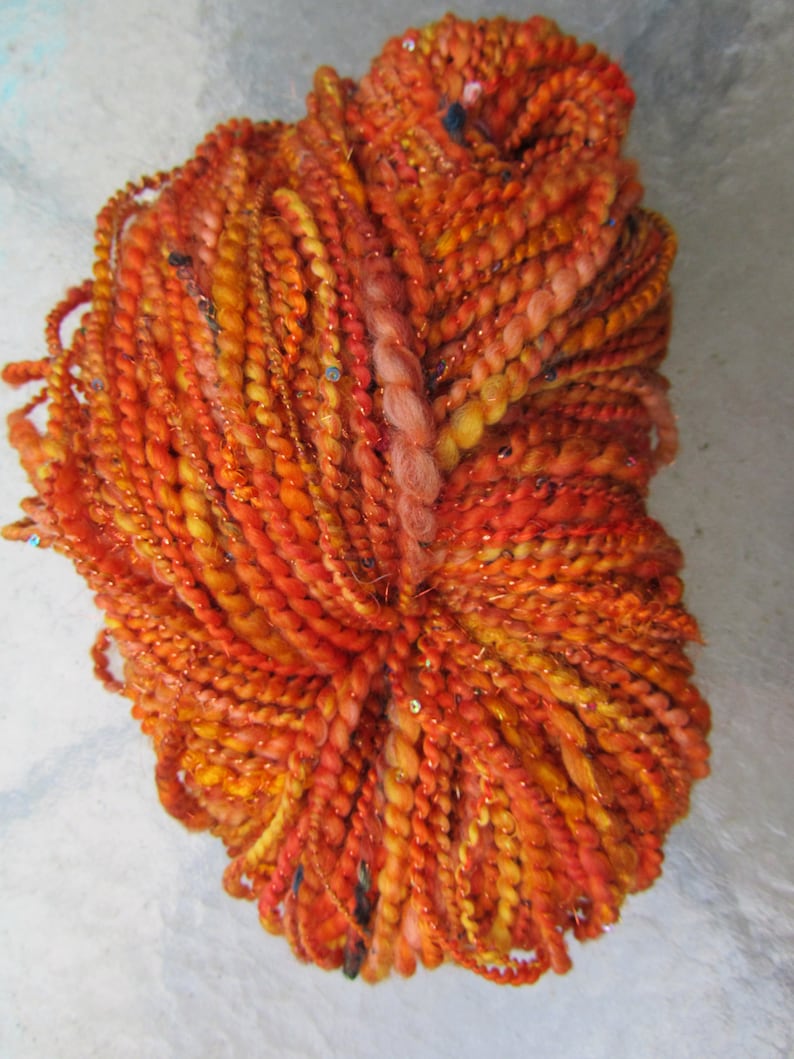 May include: A skein of textured art yarn in vibrant shades of orange, red, and yellow. The yarn features a multi-ply construction with varying thicknesses, incorporating metallic threads and small reflective sequins throughout the twisted fibers.
