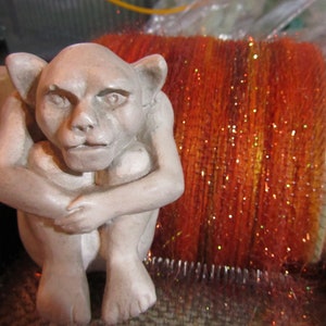 May include: A light gray, seated gargoyle figurine with pointed ears and a hunched posture, positioned in front of a large, cylindrical drum carder tool filled with vibrant, shimmering orange and red fiber with metallic gold flecks.